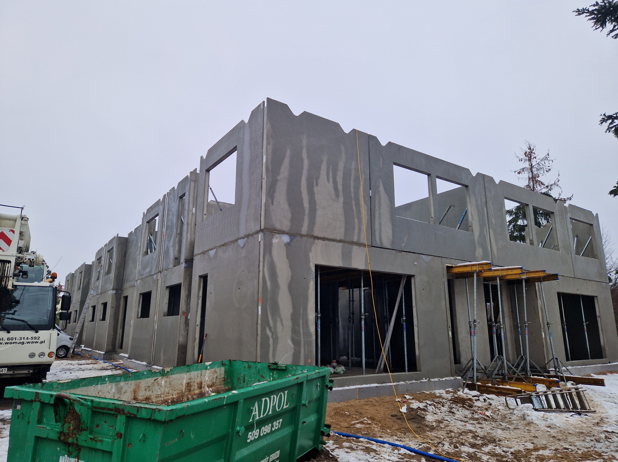 Surowy stan budynku wielorodzinnego z prefabrykatów betonowych w zimowej scenerii. Widoczne szalunki i kontener na gruz.