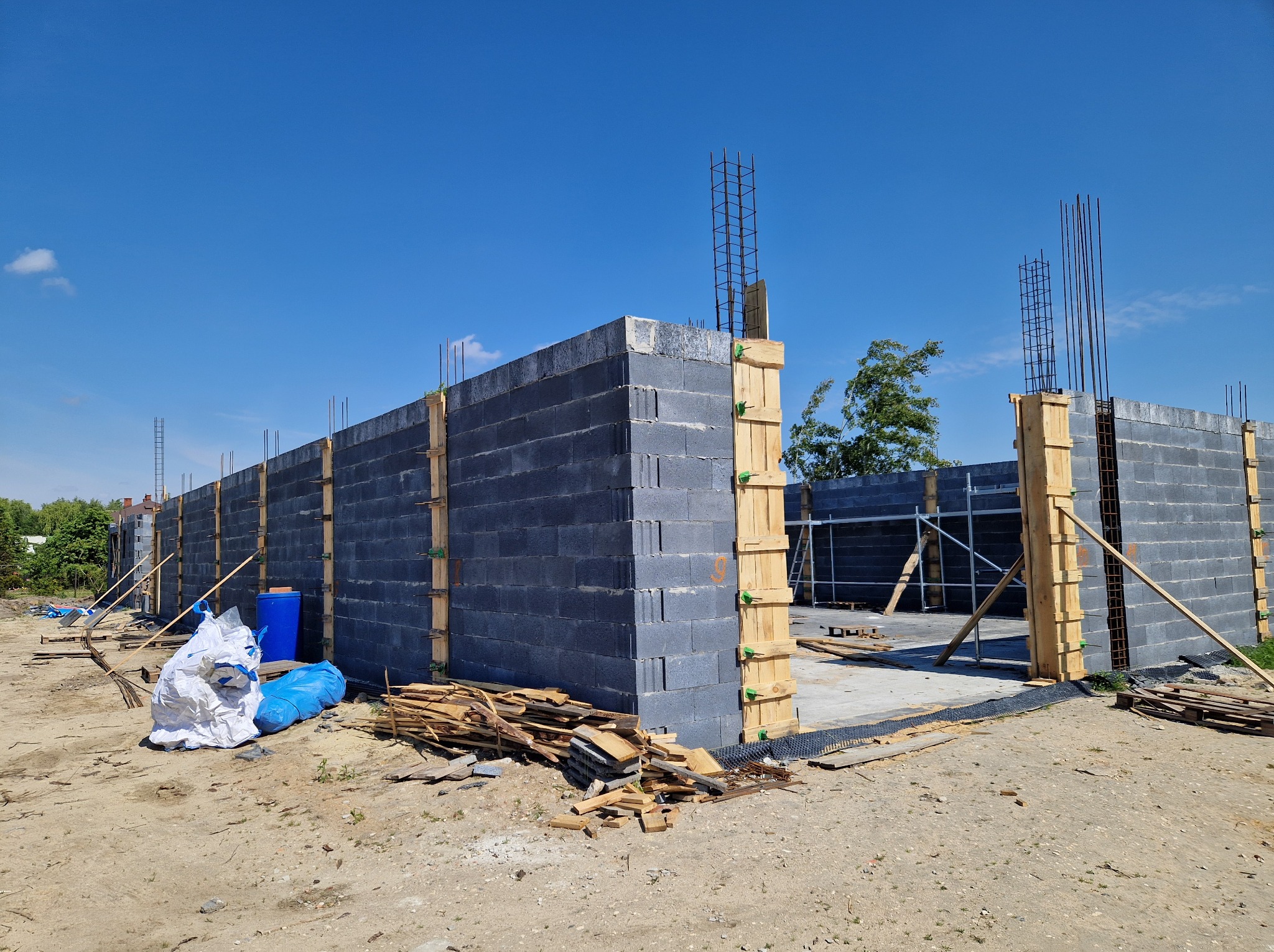 Budowa domu z bloczków betonowych w trakcie realizacji. Widoczne zbrojenia, szalunki i materiały budowlane. Błękitne niebo w tle. Świeże spojrzenie na etap budowy.