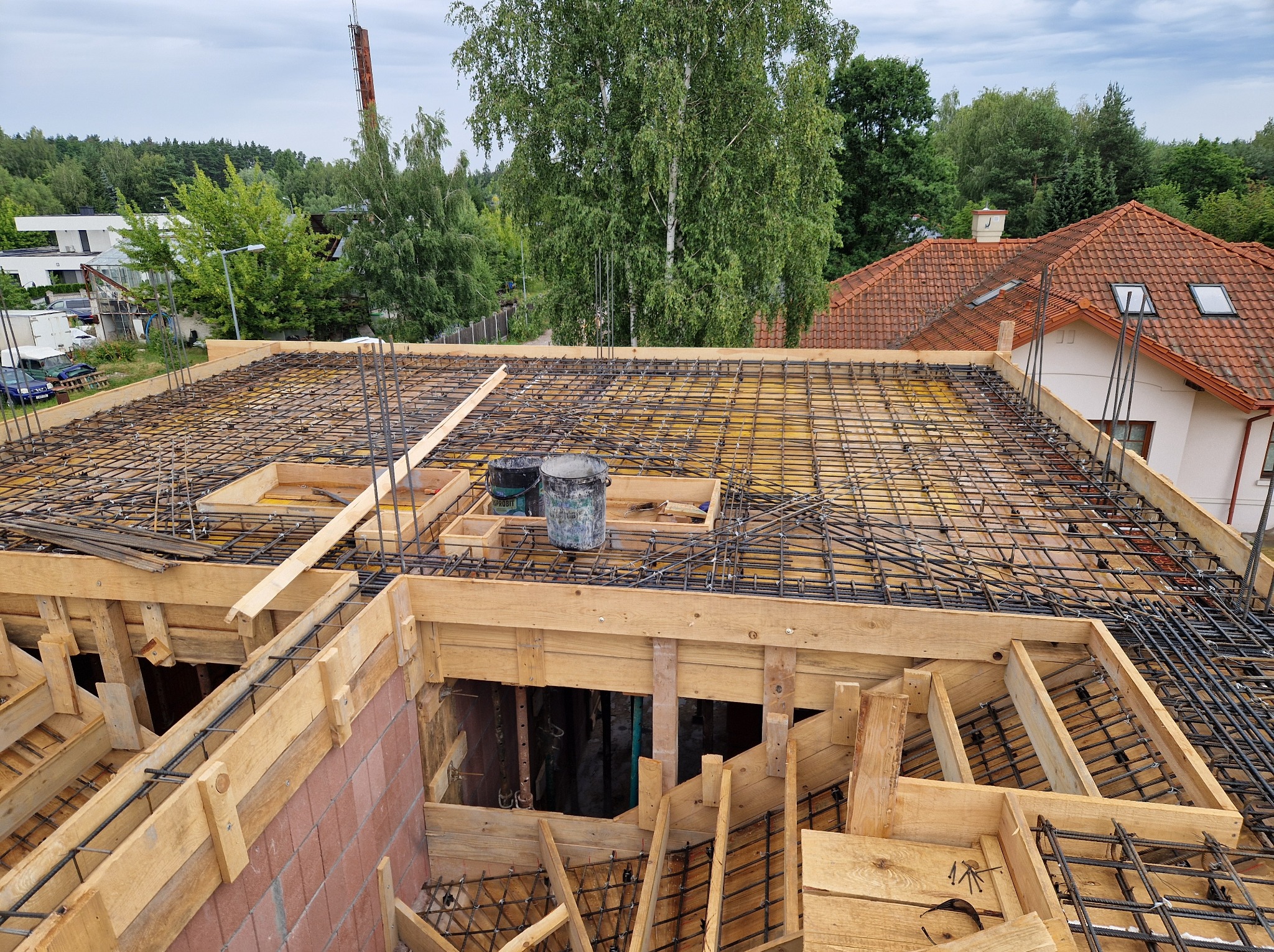 Zbrojenie stropu budynku w trakcie budowy. Widoczne drewniane szalunki, siatka zbrojeniowa i wiadra z zaprawą. W tle zabudowa jednorodzinna i zieleń.