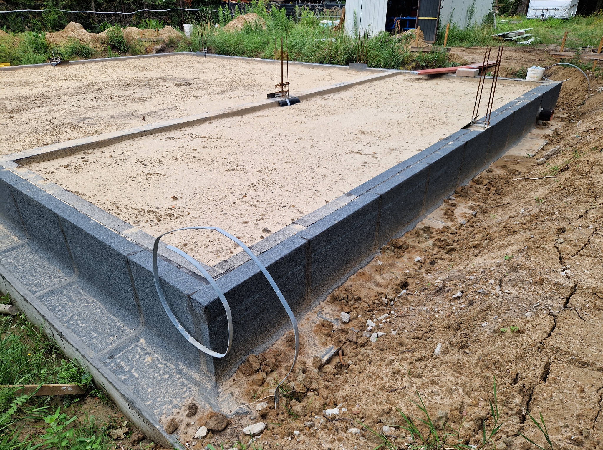 Fundament domu z izolacją termiczną i zbrojeniem pionowym, wypełniony piaskiem. Widoczne elementy szalunku i otaczający teren budowy.