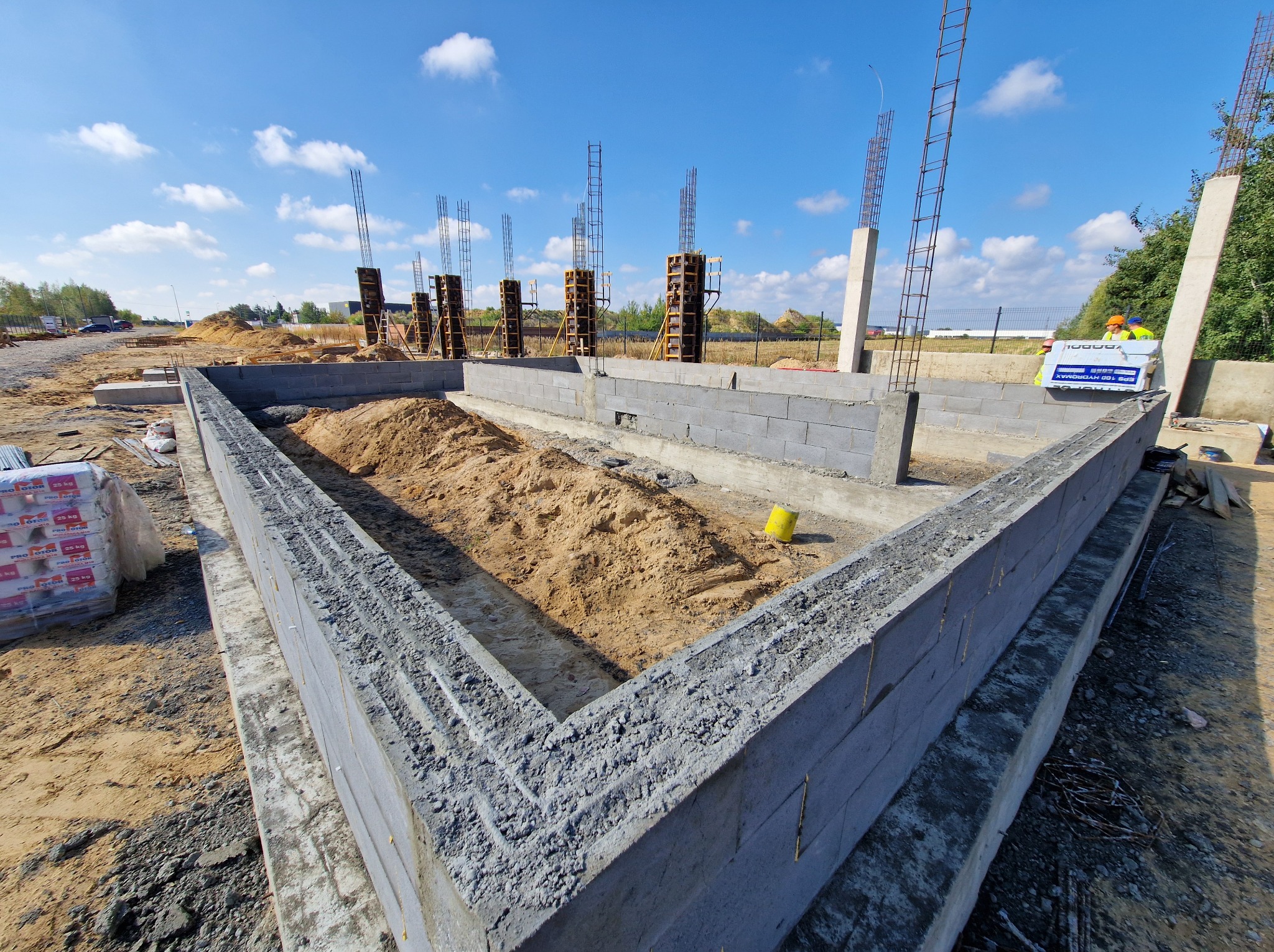 Fundamenty domu jednorodzinnego z betonu komórkowego, zbrojenia pionowe, szalunki i hałda piasku wewnątrz, błękitne niebo w tle. Etap budowy w słoneczny dzień.