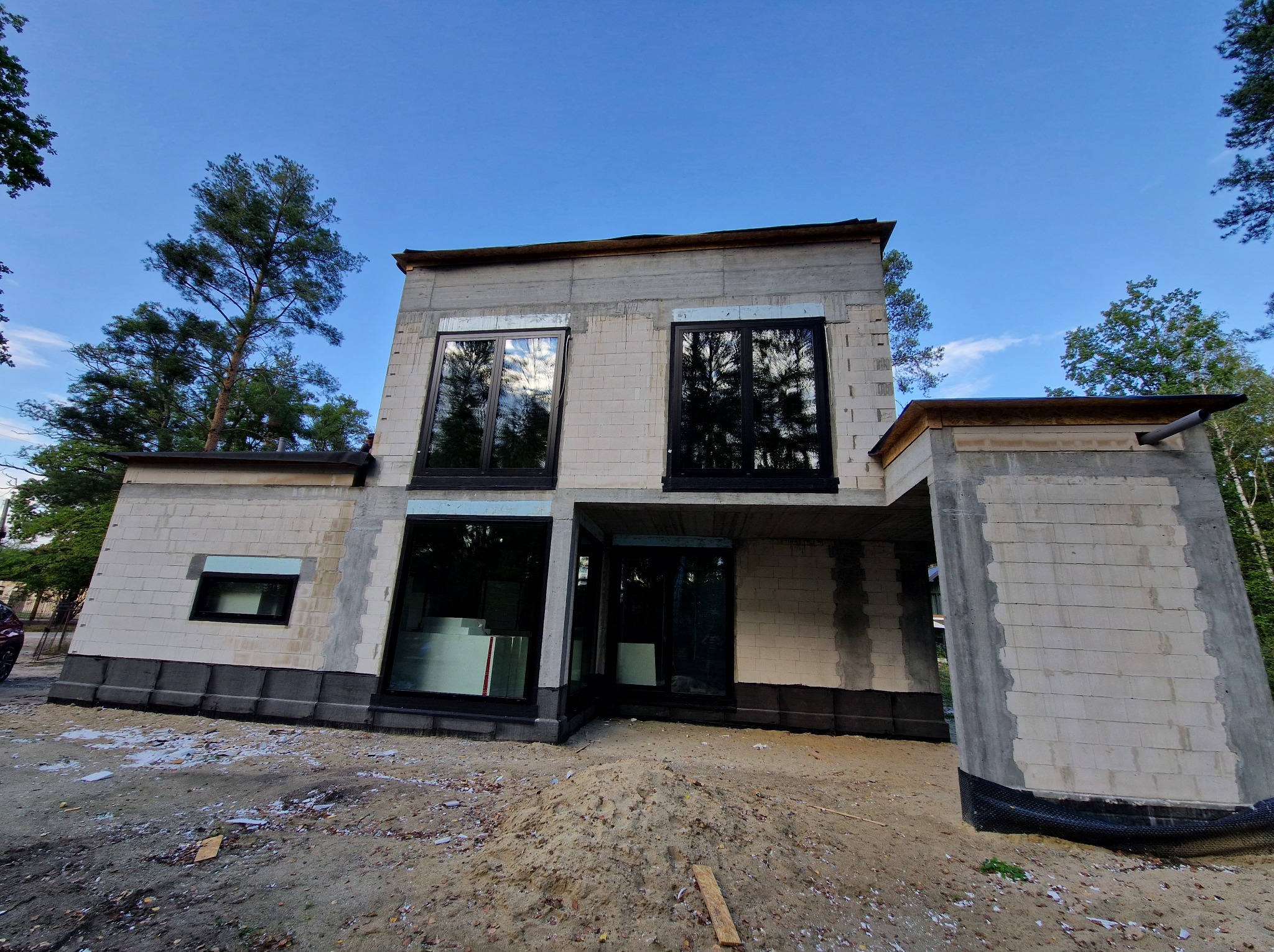 Surowy stan nowoczesnego domu z betonu komórkowego, z dużymi oknami i widoczną konstrukcją, otoczonego drzewami. W tle błękitne niebo.