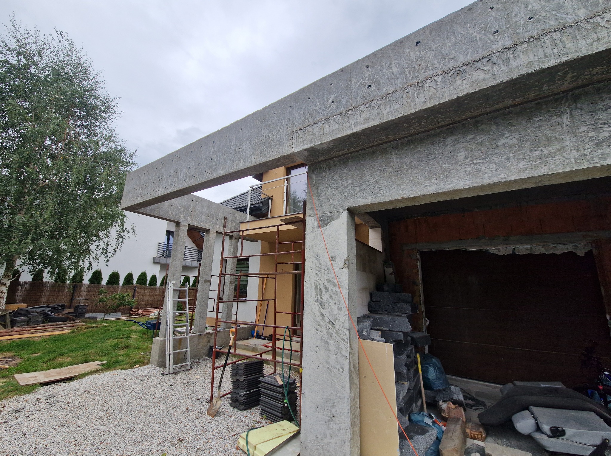 Surowa konstrukcja garażu z betonu komórkowego, z widocznym szalunkiem i rusztowaniem, na tle domu jednorodzinnego w pochmurny dzień. Etap budowy.