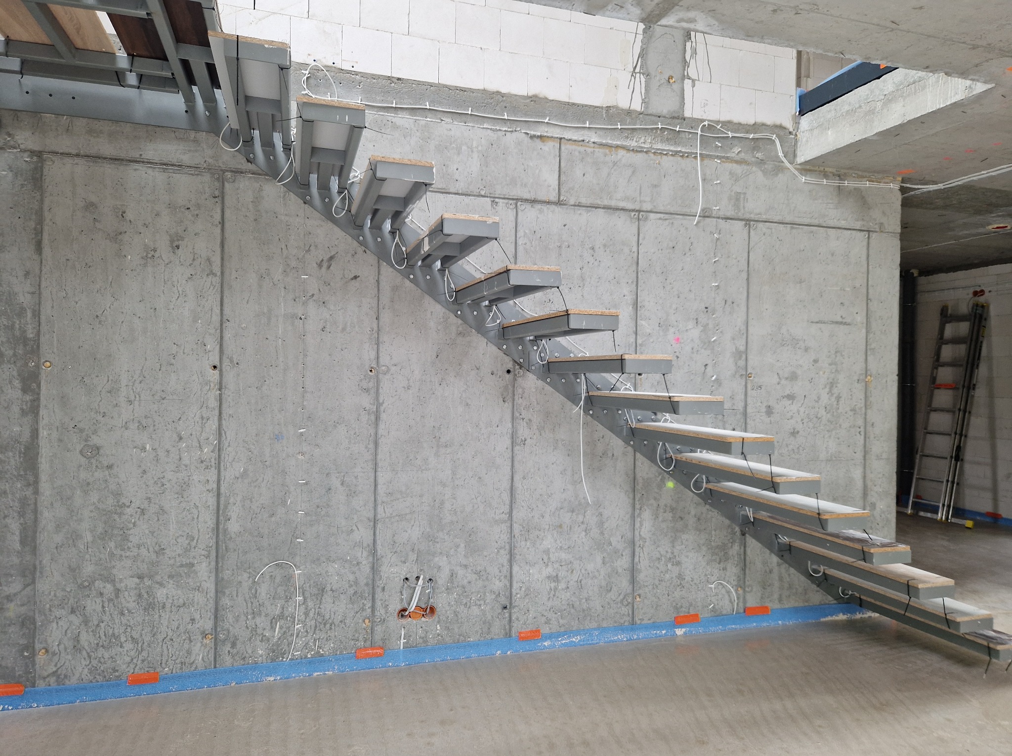 Montaż schodów stalowych z drewnianymi stopniami na surowej ścianie betonowej w trakcie prac wykończeniowych. Widoczne przewody elektryczne i drabina.