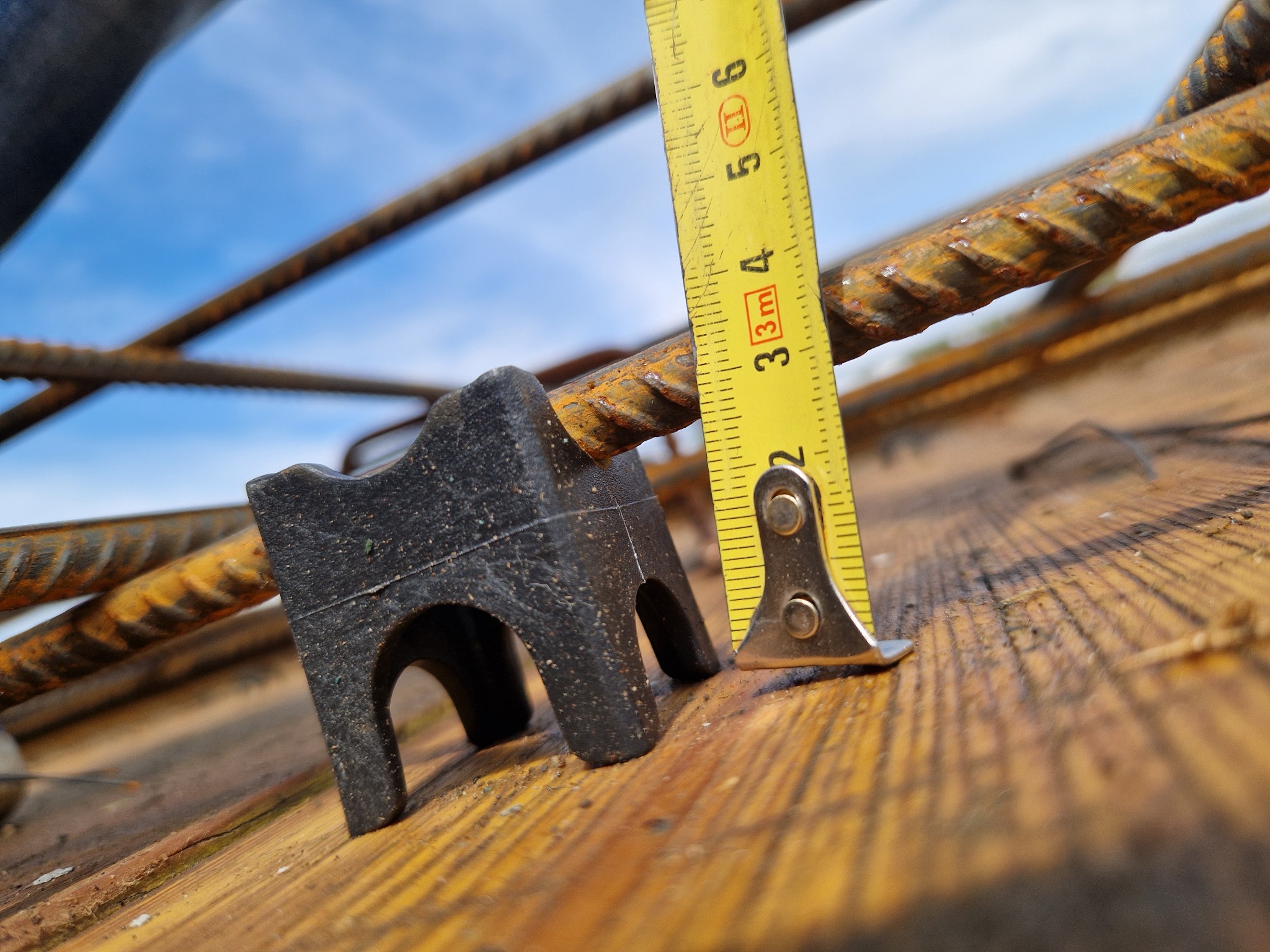 Pomiar odległości prętów zbrojeniowych za pomocą miarki, z użyciem dystansu z tworzywa. Widok z dołu na niebieskie niebo.