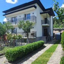 Dwupiętrowy dom z białym tynkiem i czarnym dachem, balkonami z czarnymi balustradami, widok z dołu na tle błękitnego nieba z chmurami, ozdobne krzewy przed domem.