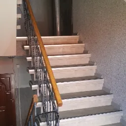 Klatka schodowa w budynku z jasnymi schodami, drewnianą poręczą i szarą, ziarnistą ścianą. W tle widoczne drzwi wejściowe i pionowa rura.