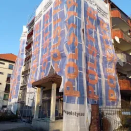 Budynek mieszkalny w trakcie ocieplania, osłonięty siatką z reklamą producenta styropianu, widoczne rusztowania i fragment wejścia do budynku.
