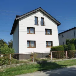 Dwupiętrowy dom z białą elewacją i brązowymi oknami, widziany z ulicy za metalową siatką ogrodzeniową, w słoneczny dzień.