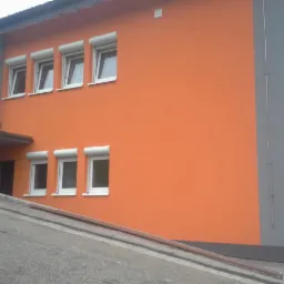 Pomarańczowa elewacja budynku z szarymi elementami, okna z roletami zewnętrznymi. Widok z dołu, podkreślający kolor i geometrię budynku.