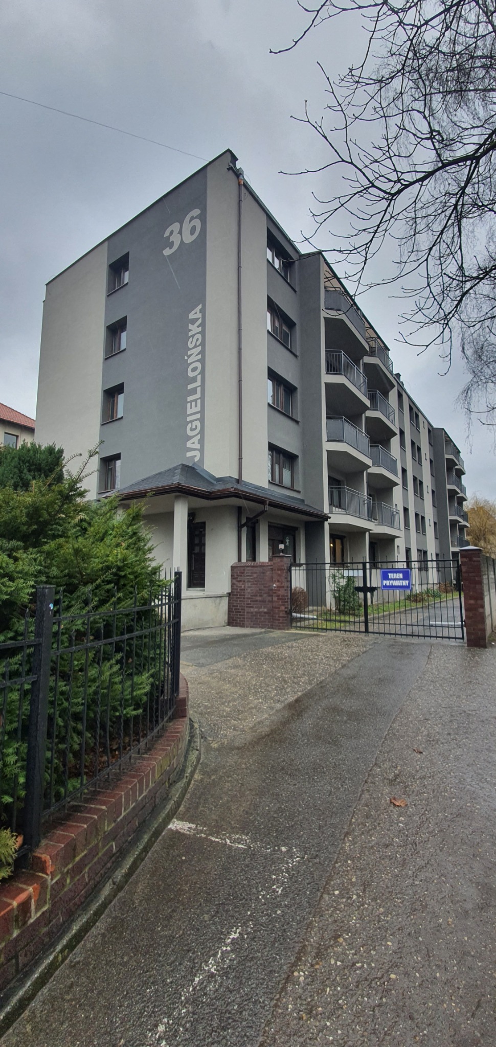 Wielopiętrowy budynek mieszkalny z szarą elewacją i białym napisem 'JAGIELLOŃSKA 36', widoczny od strony wjazdu z bramą i znakiem 'Teren prywatny'.