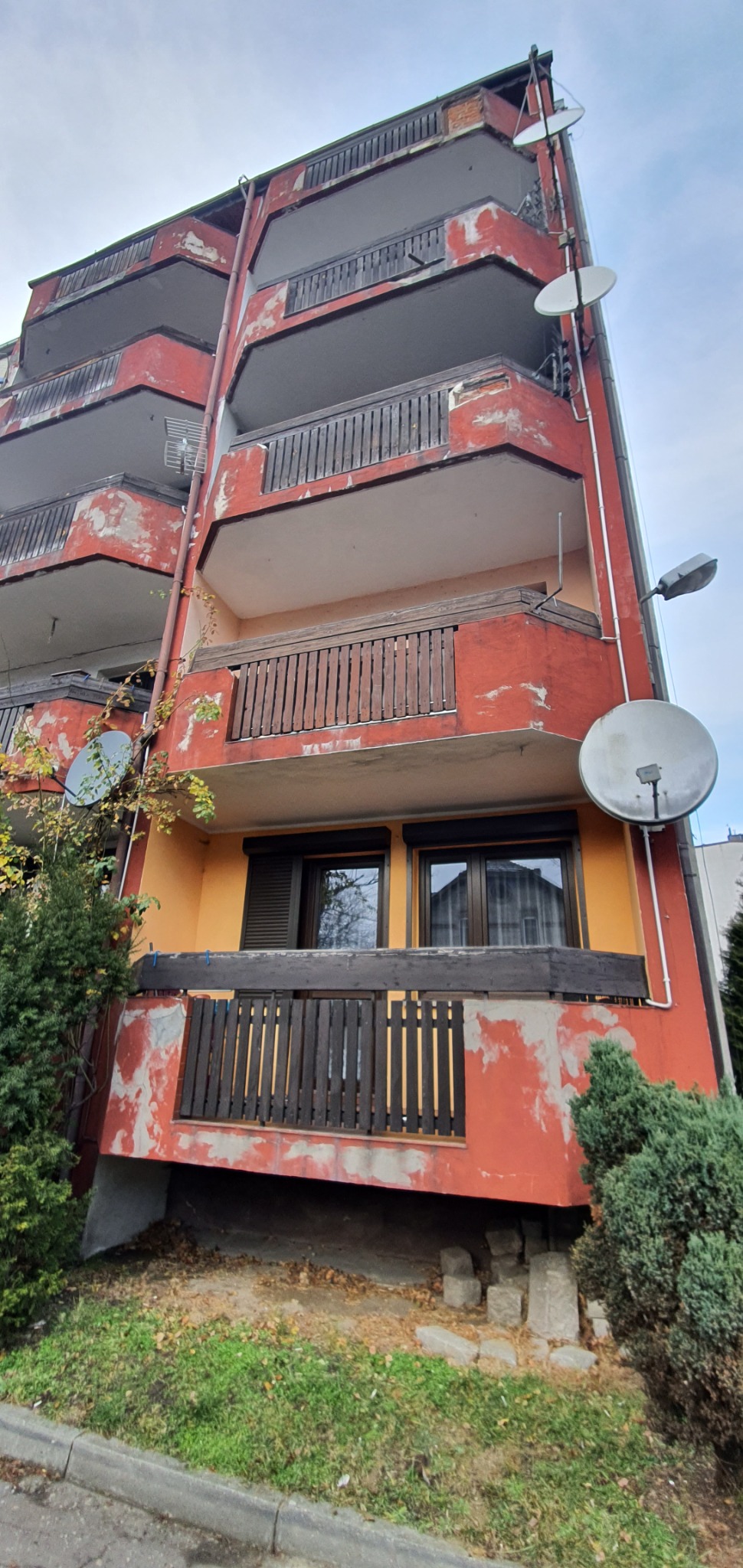 Wielopiętrowy budynek z balkonami, z widocznymi śladami łuszczącej się farby i zamontowanymi antenami satelitarnymi, sfotografowany z dołu.