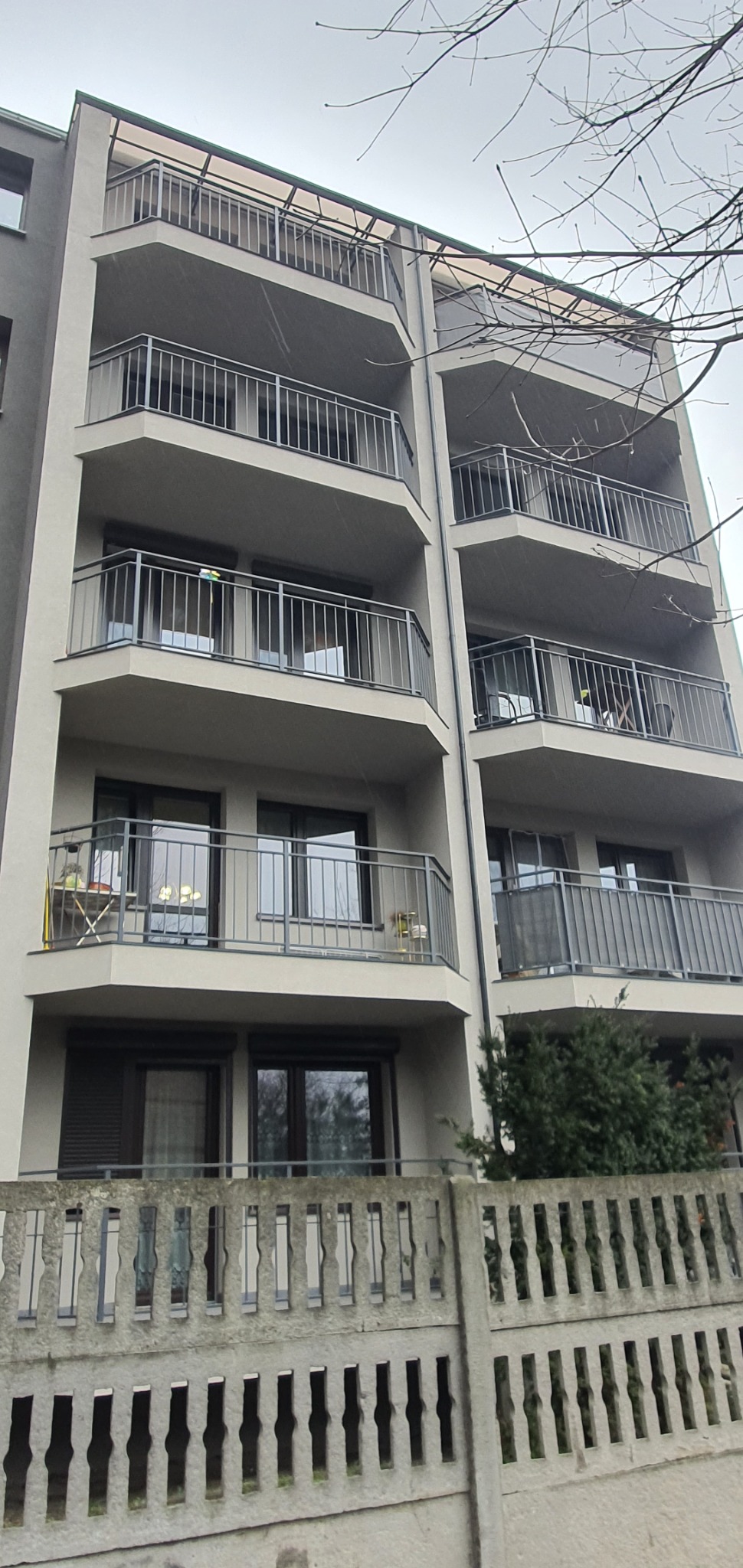 Szary budynek wielorodzinny z balkonami, widok z dołu przez betonowe ogrodzenie, na jednym z balkonów stolik i krzesła.