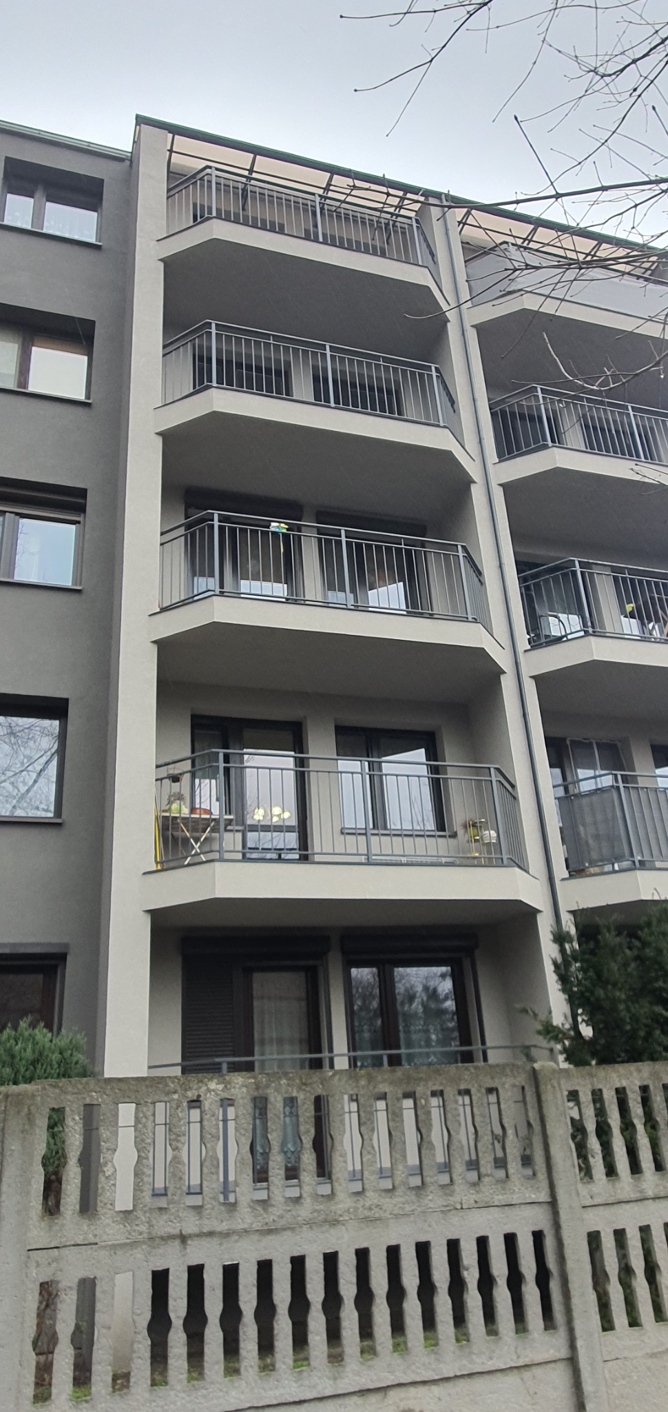 Wielopiętrowy budynek z balkonami o szarej elewacji, widoczne zza betonowego ogrodzenia, na jednym z balkonów stoi mały stolik z krzesłem.