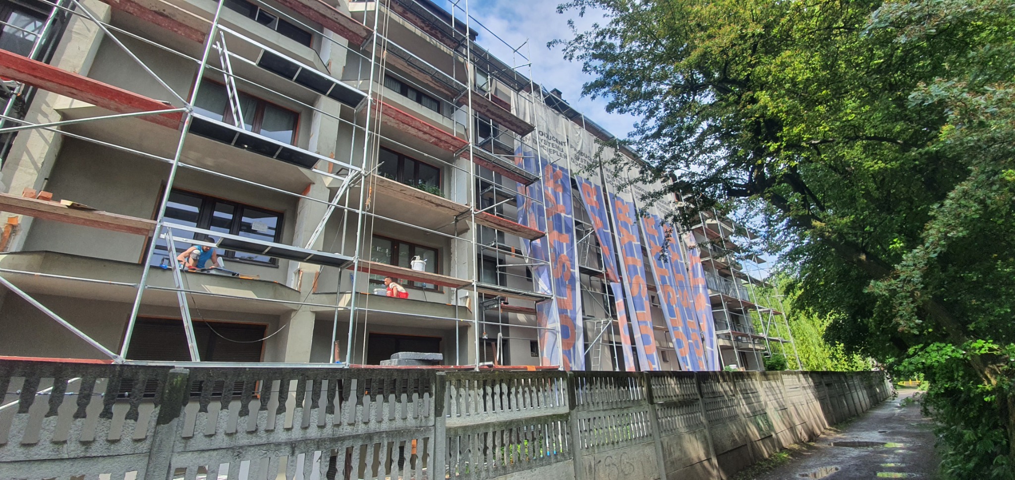 Wielopiętrowy budynek w trakcie prac elewacyjnych, osłonięty banerami reklamowymi, z widocznymi rusztowaniami i pracownikami na różnych poziomach, widziany zza betonowego ogrodzenia.