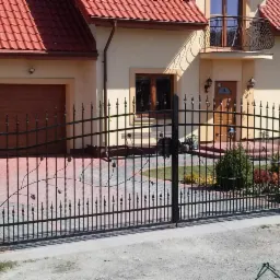 Kuta brama wjazdowa z motywem roślinnym, czarna, na tle domu z czerwoną dachówką i brukowanym podjazdem. Elementy dekoracyjne w formie liści i pąków.