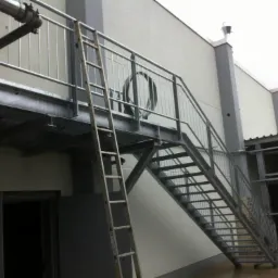 Zewnętrzne stalowe schody z podestem i drabina oparta o konstrukcję, prowadzące do wejścia w budynku o jasnej elewacji. Widoczne elementy instalacji na ścianie.