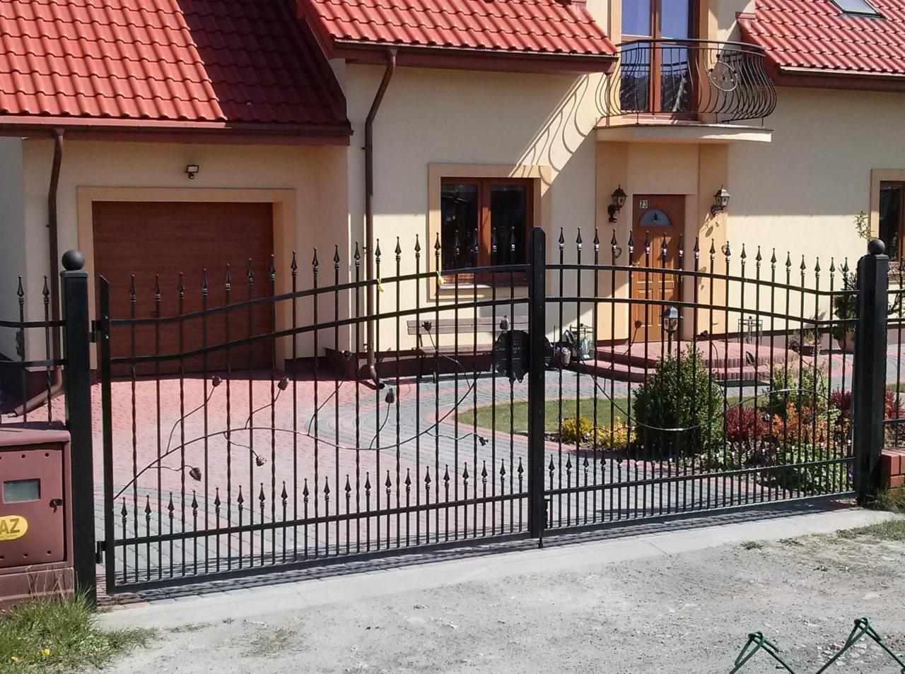 Kuta brama wjazdowa z motywem roślinnym, czarna, na tle domu z czerwoną dachówką i brukowanym podjazdem. Elementy dekoracyjne w formie liści i pąków.