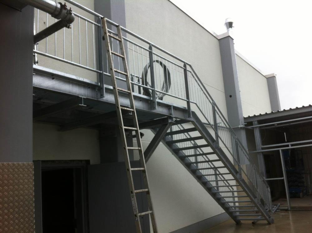 Zewnętrzne stalowe schody z podestem i drabina oparta o konstrukcję, prowadzące do wejścia w budynku o jasnej elewacji. Widoczne elementy instalacji na ścianie.