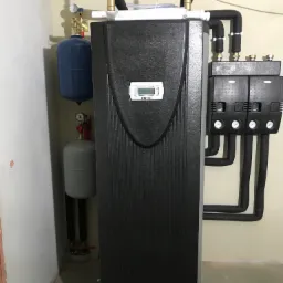 Czarna pompa ciepła Dimplex z podłączonymi rurami, manometrami i zbiornikami wyrównawczymi, zainstalowana w pomieszczeniu z beżowymi ścianami.