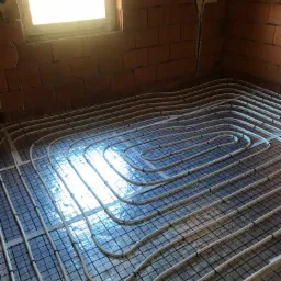 Białe rury ogrzewania podłogowego ułożone spiralnie na czarnej siatce, na podłodze z folii odblaskowej w surowym wnętrzu z ceglanymi ścianami i oknem.