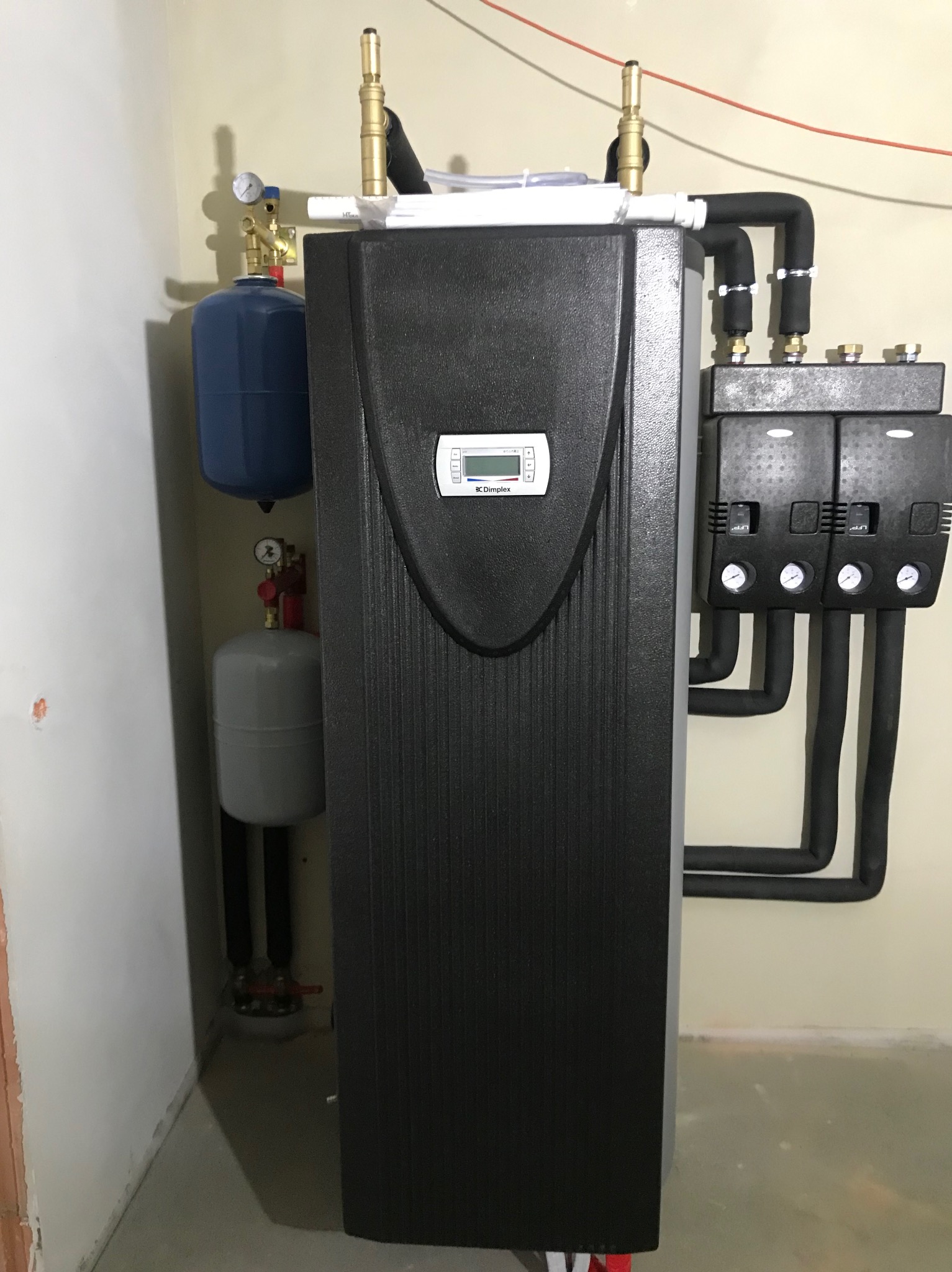 Czarna pompa ciepła Dimplex z podłączonymi rurami, manometrami i zbiornikami wyrównawczymi, zainstalowana w pomieszczeniu z beżowymi ścianami.