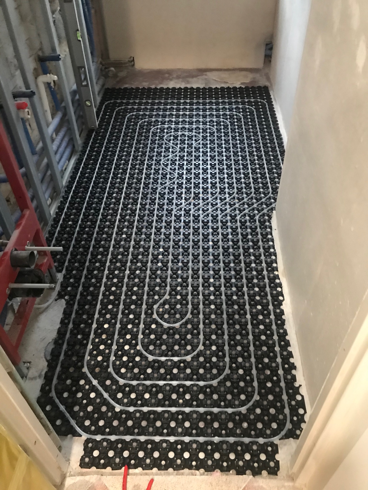Rozłożona instalacja ogrzewania podłogowego z rurkami na czarnej macie, widoczna w trakcie montażu w pomieszczeniu z surowymi ścianami i instalacjami hydraulicznymi.