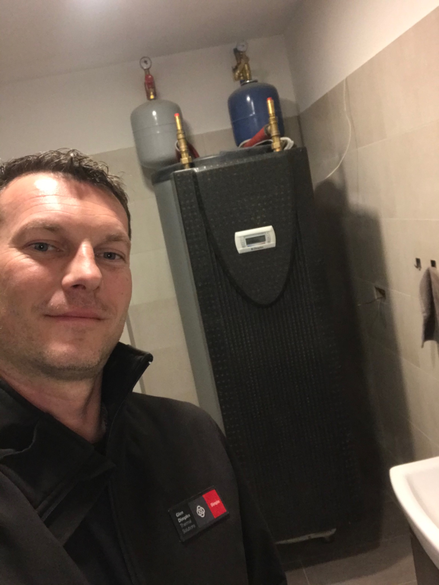 Mężczyzna robi selfie na tle zamontowanego zbiornika buforowego z dwoma naczyniami wzbiorczymi w łazience z szarymi płytkami.