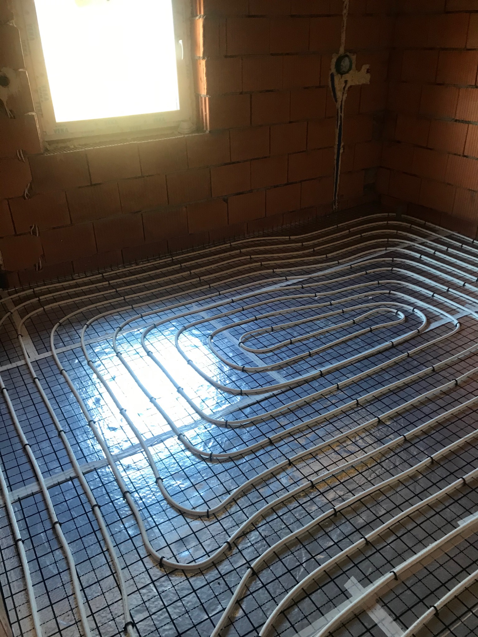 Białe rury ogrzewania podłogowego ułożone spiralnie na czarnej siatce, na podłodze z folii odblaskowej w surowym wnętrzu z ceglanymi ścianami i oknem.