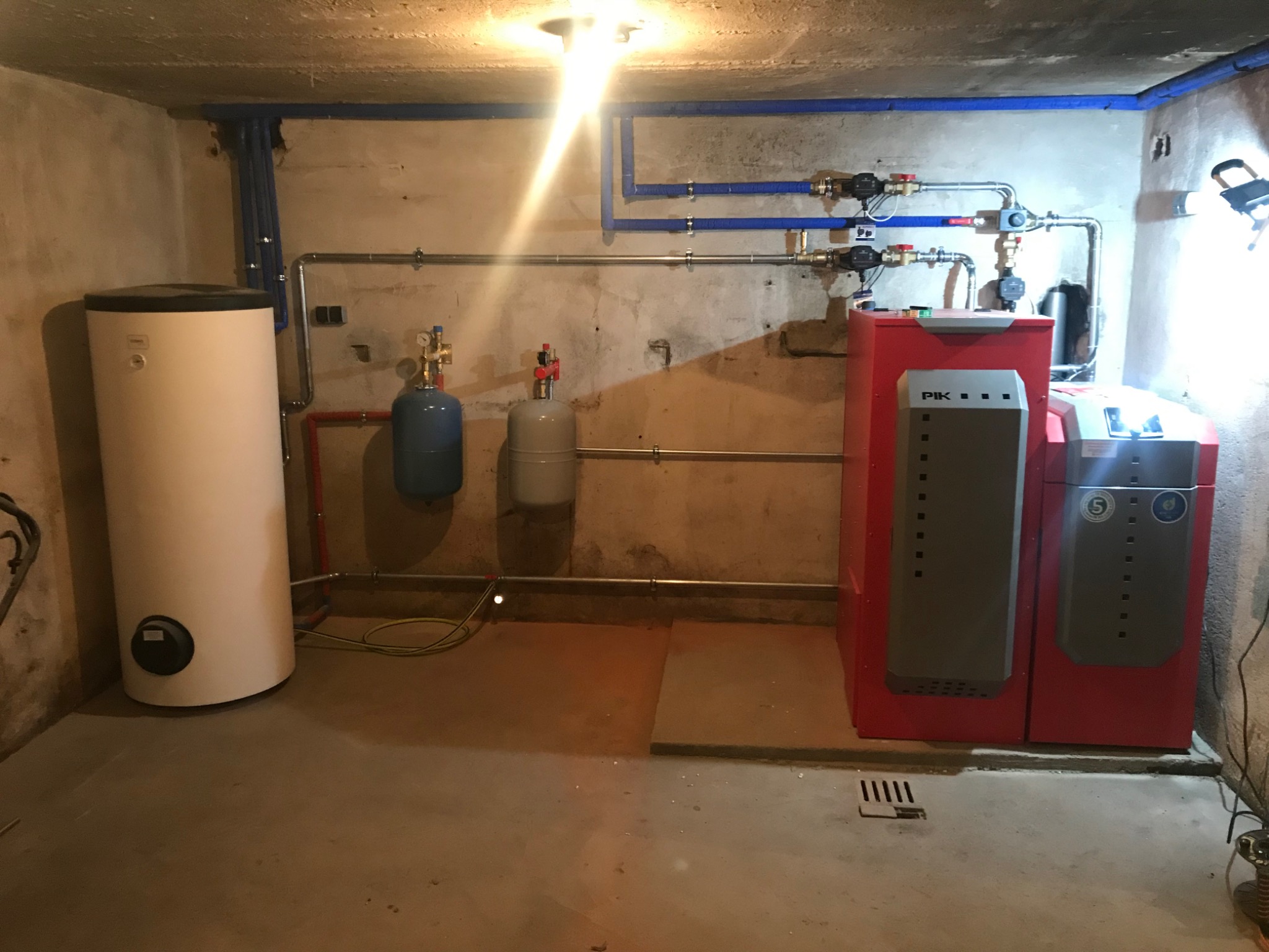 Instalacja grzewcza w piwnicy: czerwony piec z szarymi panelami, biały zasobnik ciepłej wody, niebieskie rury na ścianie, zbiorniki wyrównawcze i metalowe rury. Widoczna lampa halogenowa...