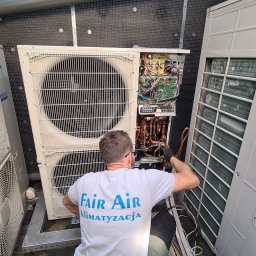 klimatyzacjewarszawa.pl - Technik w okularach, w koszulce firmy Fair Air, serwisuje otwartą jednostkę zewnętrzną klimatyzacji, z widocznymi przewodami i elektroniką.