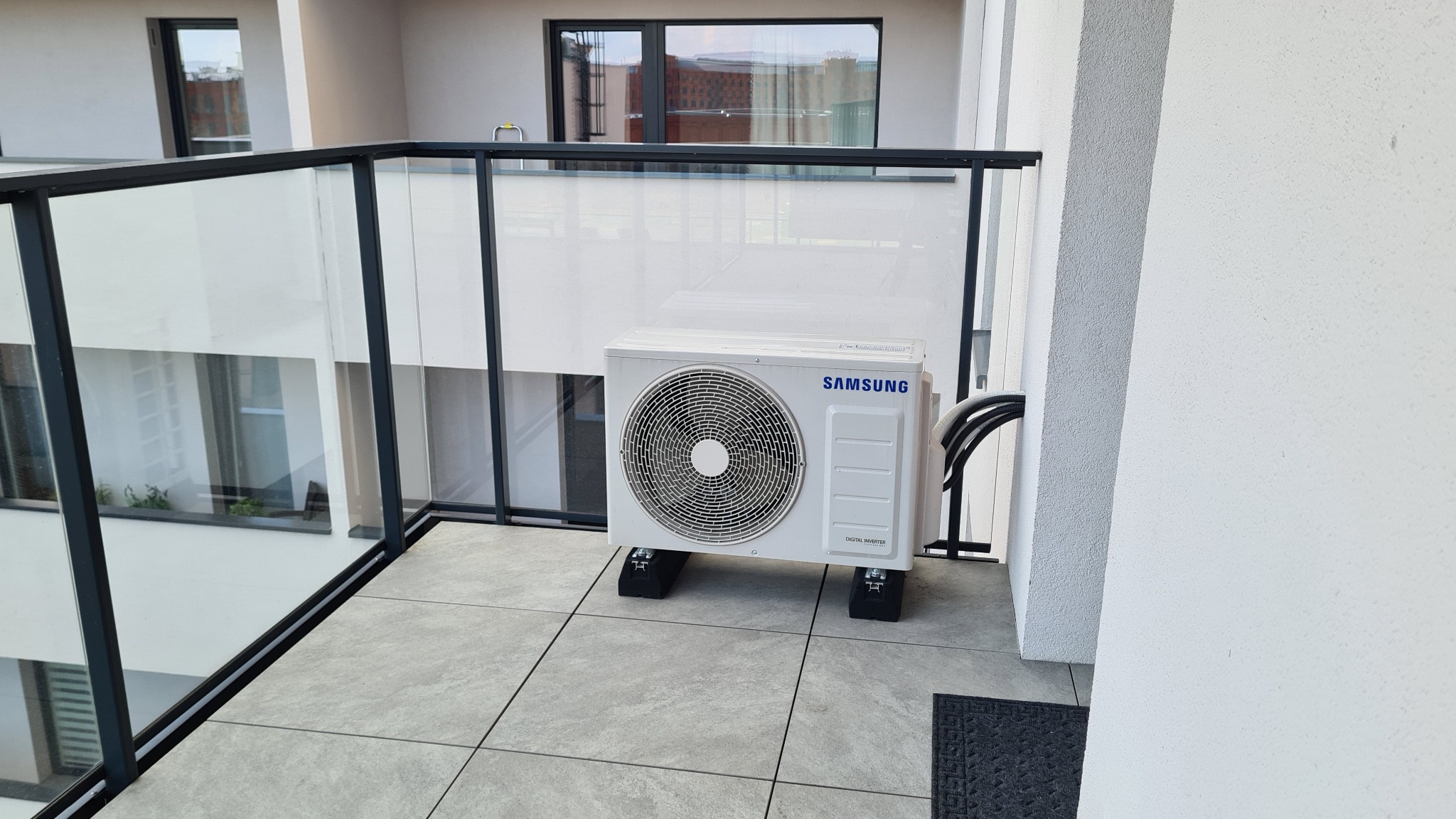 Biała jednostka zewnętrzna klimatyzacji Samsung Digital Inverter zamontowana na czarnych podkładkach antywibracyjnych na balkonie z szklaną balustradą i szarymi płytkami podłogowymi.