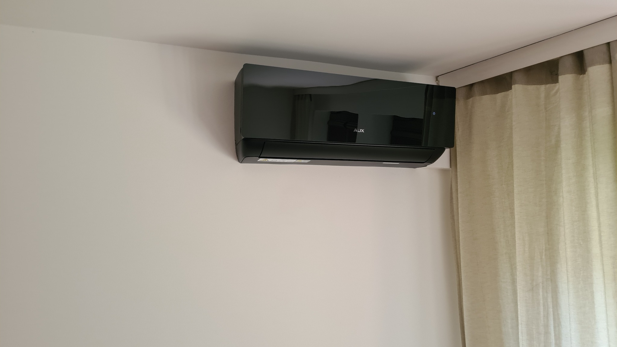 Czarny klimatyzator marki AUX zamontowany na białej ścianie w rogu pokoju, obok zasłony w kolorze ecru.