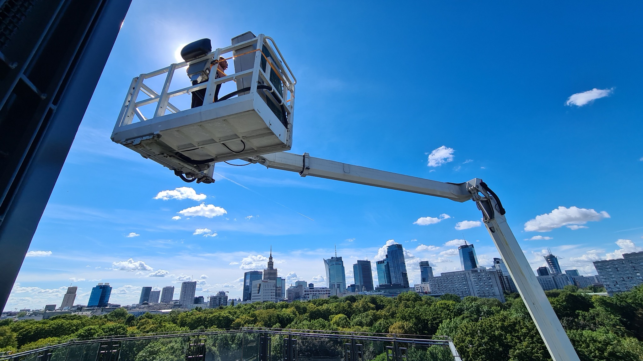 Podnośnik koszowy z pracownikiem na tle panoramy Warszawy i błękitnego nieba z chmurami, widok z góry.