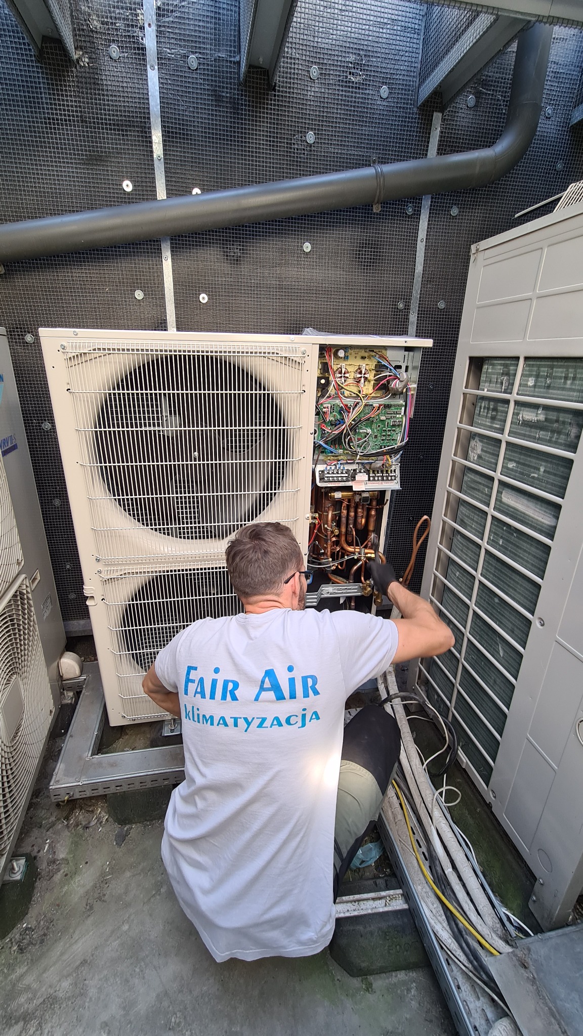 Technik w okularach, w koszulce firmy Fair Air, serwisuje otwartą jednostkę zewnętrzną klimatyzacji, z widocznymi przewodami i elektroniką.