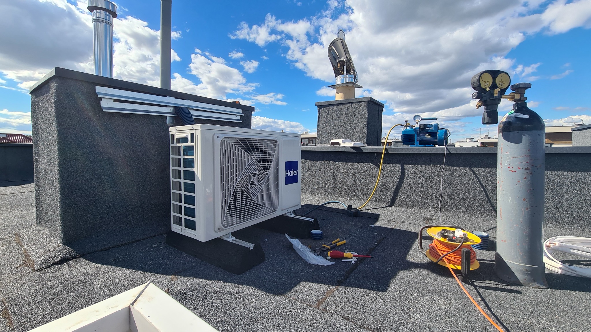 Biała jednostka zewnętrzna klimatyzacji Haier zamontowana na płaskim dachu, obok butla z gazem, pompa próżniowa i zwinięty przedłużacz. Widoczne narzędzia i elementy instalacji.
