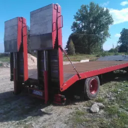 Czerwona, dwuosiowa przyczepa niskopodwoziowa z najazdami hydraulicznymi, zaparkowana na placu z widocznymi stertami ziemi w tle.