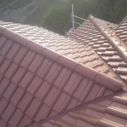 Czerwona, ceramiczna dachówka karpiówka na połaci dachu. Widoczny system mocowań i fragment rusztowania w tle, w słoneczny dzień.