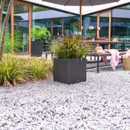 Nowoczesny ogród z szarym żwirem, kwadratowe donice z ozdobnymi trawami, drewniany stół z parasolem i dekoracjami w tle.