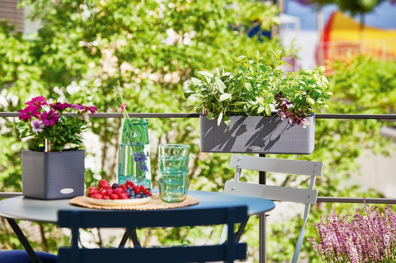 Balkon z metalową barierką, na której zawieszono donice z ziołami. Na stoliku o metalowej konstrukcji stoją kwiaty w doniczce, dzbanek z wodą i talerz z owocami.