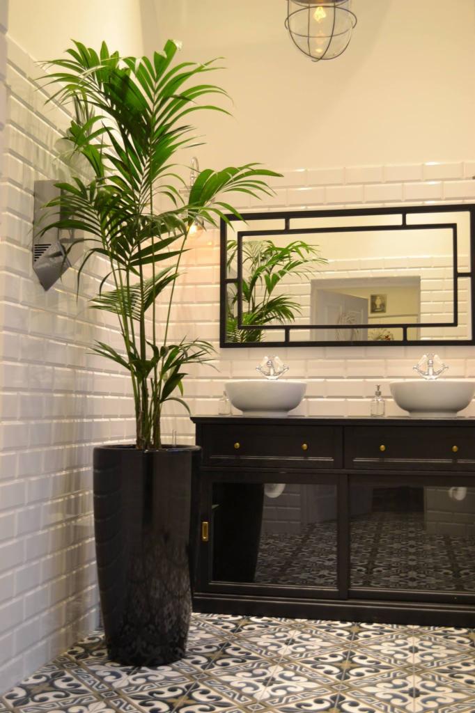 Wnętrze łazienki z dużym kwiatem doniczkowym w czarnej, lakierowanej donicy, umywalki nablatowe, lustro w czarnej ramie, białe płytki na ścianach i wzorzysta podłoga.