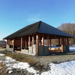 Drewniana konstrukcja altany z czarnym dachem, częściowo wykończona kamieniem, na tle zimowego krajobrazu z resztkami śniegu i lasem w oddali, sfotografowana w jasnym, słonecznym świetle.