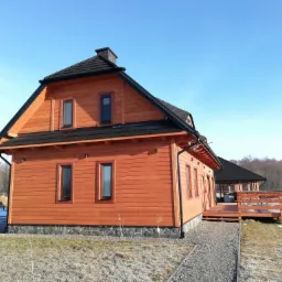 Nowoczesny, dwukondygnacyjny dom z bali o pomarańczowej elewacji, z czarnym dachem i kamienną podmurówką, z widocznym fragmentem tarasu i altany w tle, sfotografowany w słoneczny dzień.