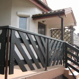 Czarna, drewniana balustrada zewnętrzna z ukośnymi szczeblami, mocowana na gwoździe, prowadząca do wejścia domu z widoczną kratką na werandzie w tle.