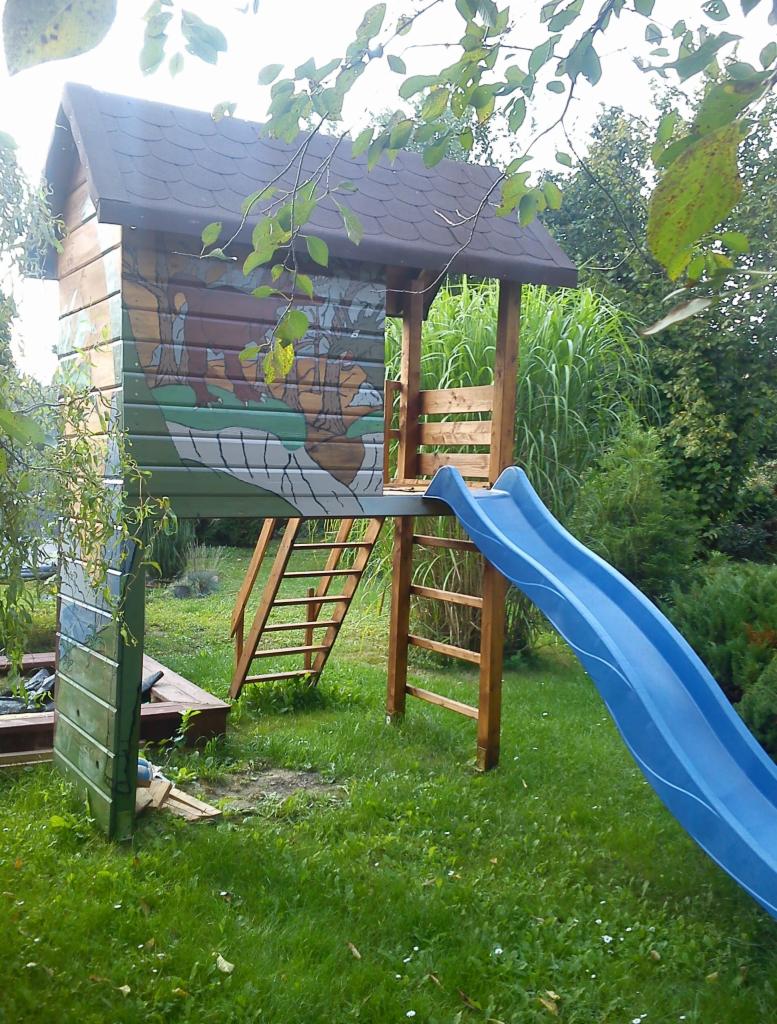 Drewniany domek dla dzieci ze zjeżdżalnią, pomalowany w motywy leśne, umieszczony w ogrodzie z bujną zielenią.