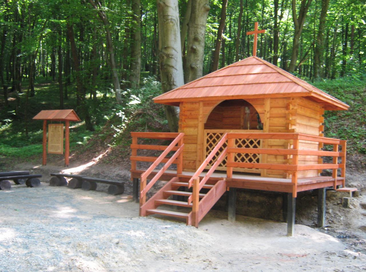 Drewniana kapliczka z krzyżem na dachu, zbudowana z bali, z ażurowymi drzwiczkami i schodami, umiejscowiona w lesie, obok widoczna tablica informacyjna i ławki wykonane z pni drzew.