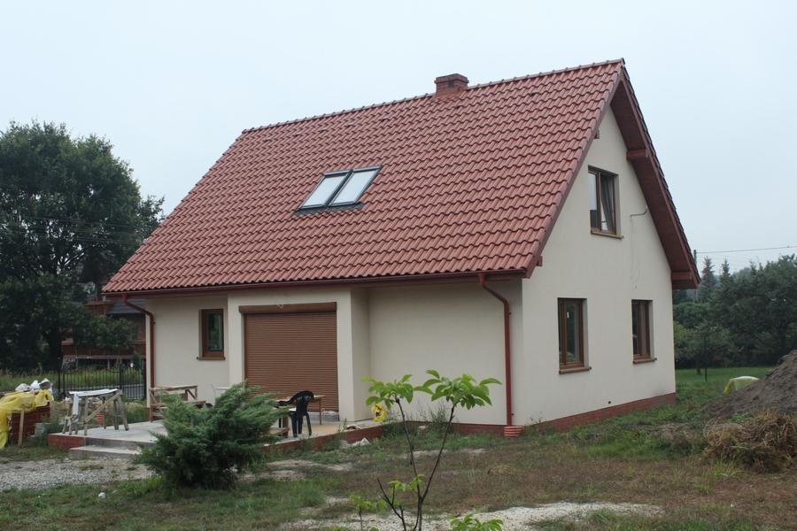 Gotowy dom jednorodzinny z czerwoną dachówką, dwoma oknami dachowymi, beżową elewacją i brązowymi roletami zewnętrznymi. Widoczne elementy wykończeniowe i zagospodarowanie terenu wokół domu.