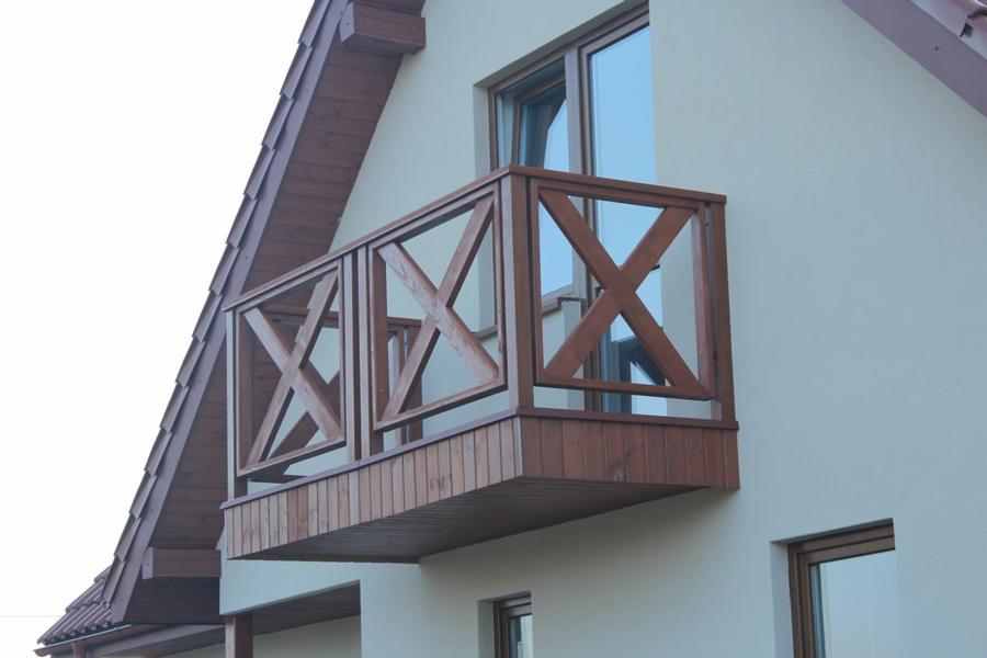 Balkon z drewnianą balustradą w kształcie krzyży, zamontowany na jasnej elewacji domu z brązowym dachem.