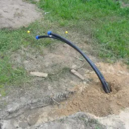 Dwie czarne rury z niebieskimi końcówkami wychodzące z wykopu w ziemi, obok trawa i drewniane kliny.