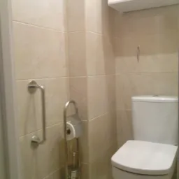 Jasna łazienka po remoncie: kremowe płytki, biała toaleta i szafka wisząca. Widoczny uchwyt dla niepełnosprawnych i stojak na papier toaletowy.